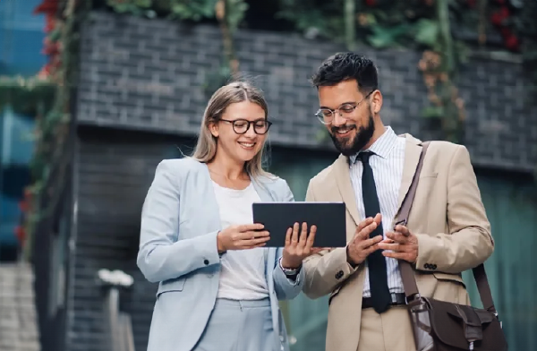 Un homme et une femme en tenue professionnelle se tiennent debout et regardent une tablette en souriant.